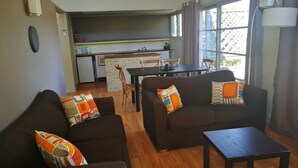 A living room with two sofas, a coffee table, and a dining table. There is a kitchen in the background with a window and curtains. - Vineyard Hill (Lovedale)