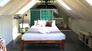 A bedroom with a sloped ceiling, a bed with pillows, a bedside table, a hanging plant, and a window. - Vineyard Hill (Lovedale)