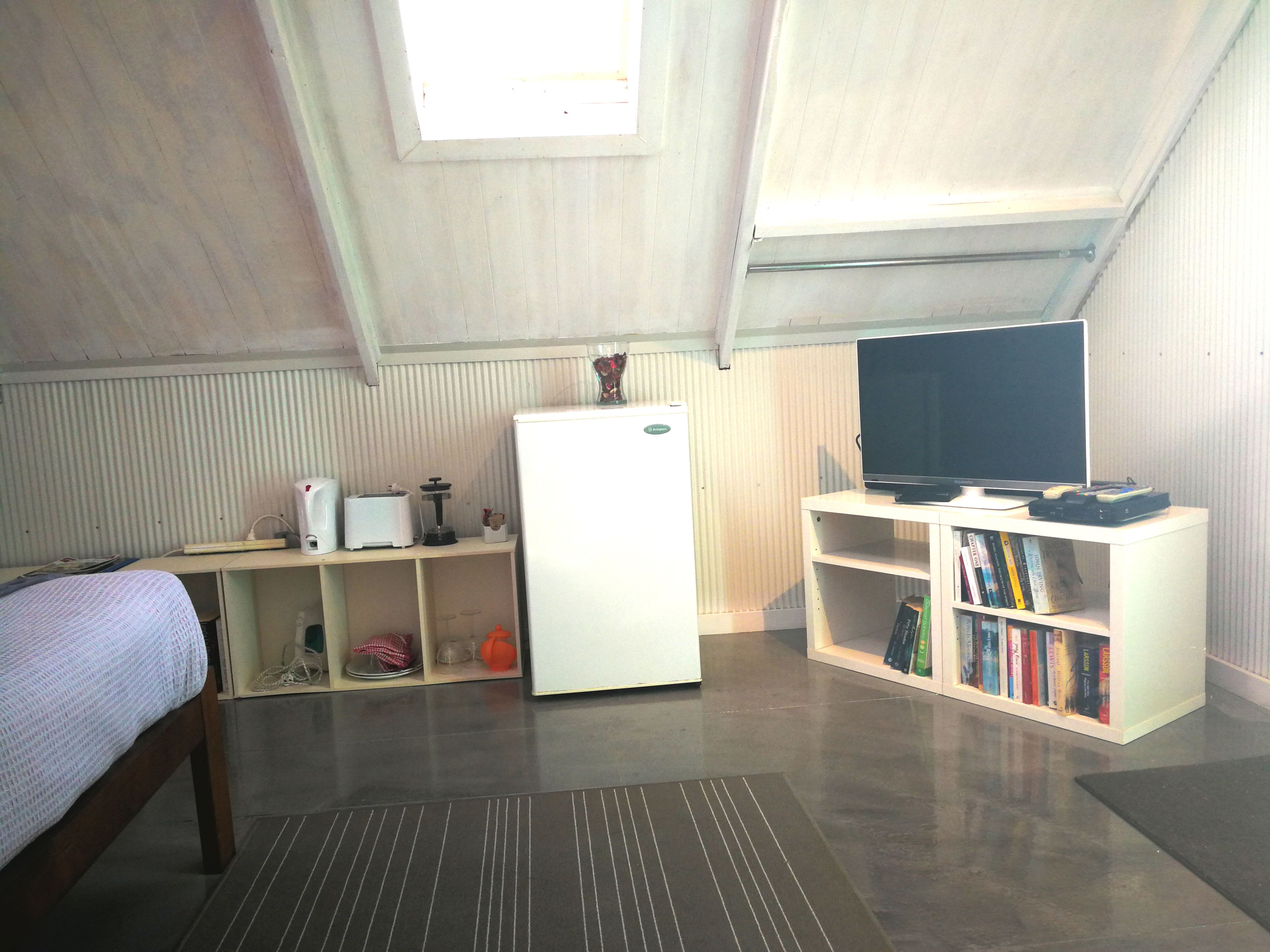 A small, well-lit room with a bed, a white refrigerator, a TV on a stand, and a bookshelf with books.