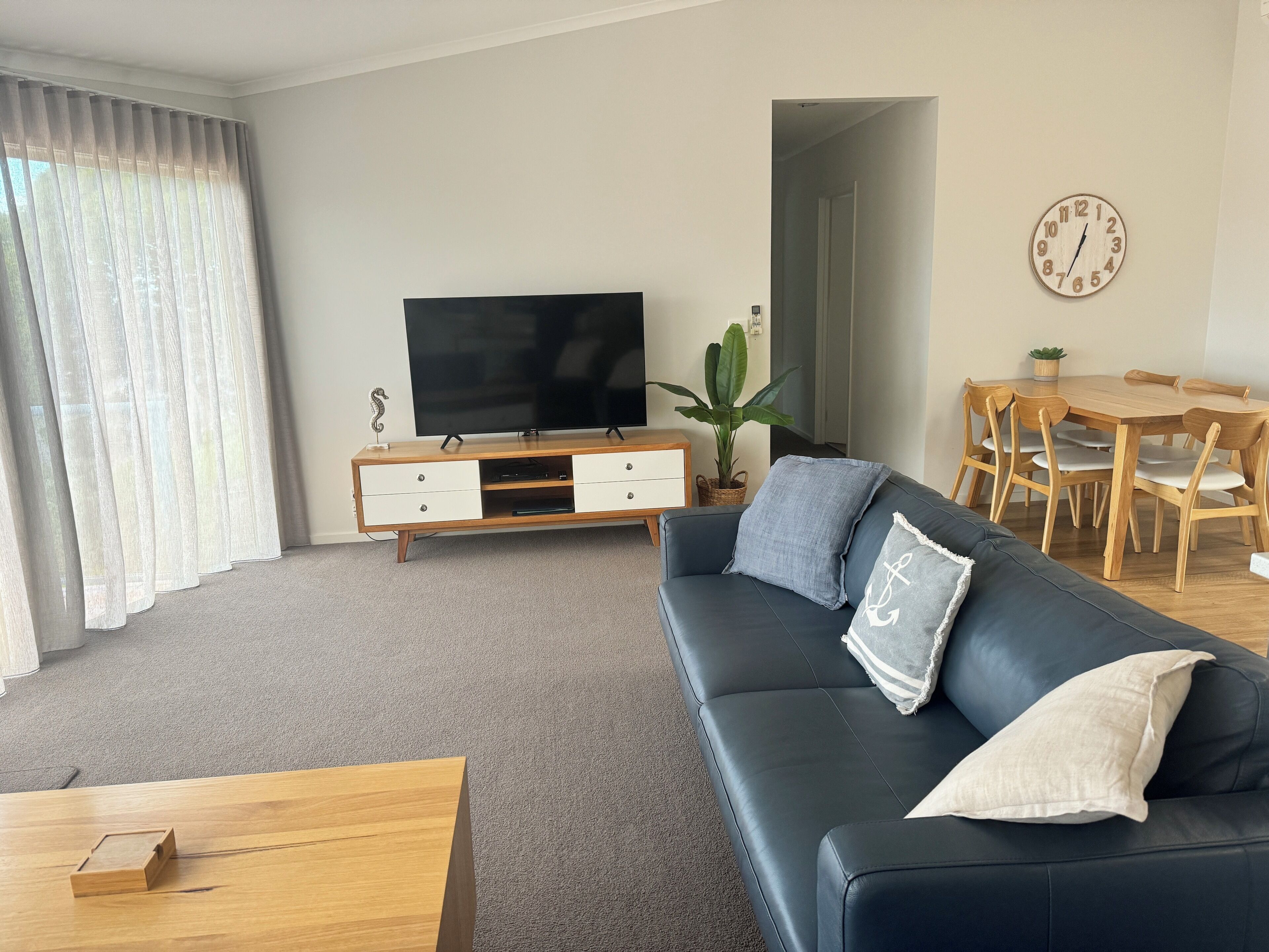 A living room with a black leather sofa, wooden coffee table, and a TV on a wooden stand. There is a dining table with chairs and a clock on the wall.