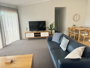 A living room with a black leather sofa, wooden coffee table, and a TV on a wooden stand. There is a dining table with chairs and a clock on the wall. - Bluewater Apartments Merimbula (Merimbula)