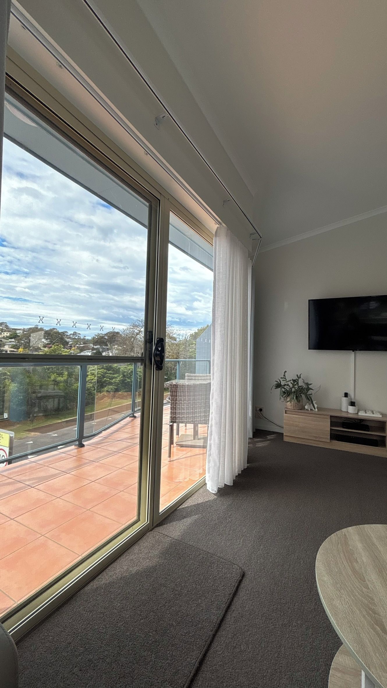 A modern living room with a large sliding glass door leading to an outdoor patio, a flat-screen TV mounted on the wall, and a minimalist furniture arrangement.