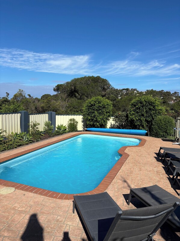 A swimming pool with a tiled edge, surrounded by a brick patio. There are lounge chairs and a blue water slide in the background. - Bluewater Apartments Merimbula (Merimbula)