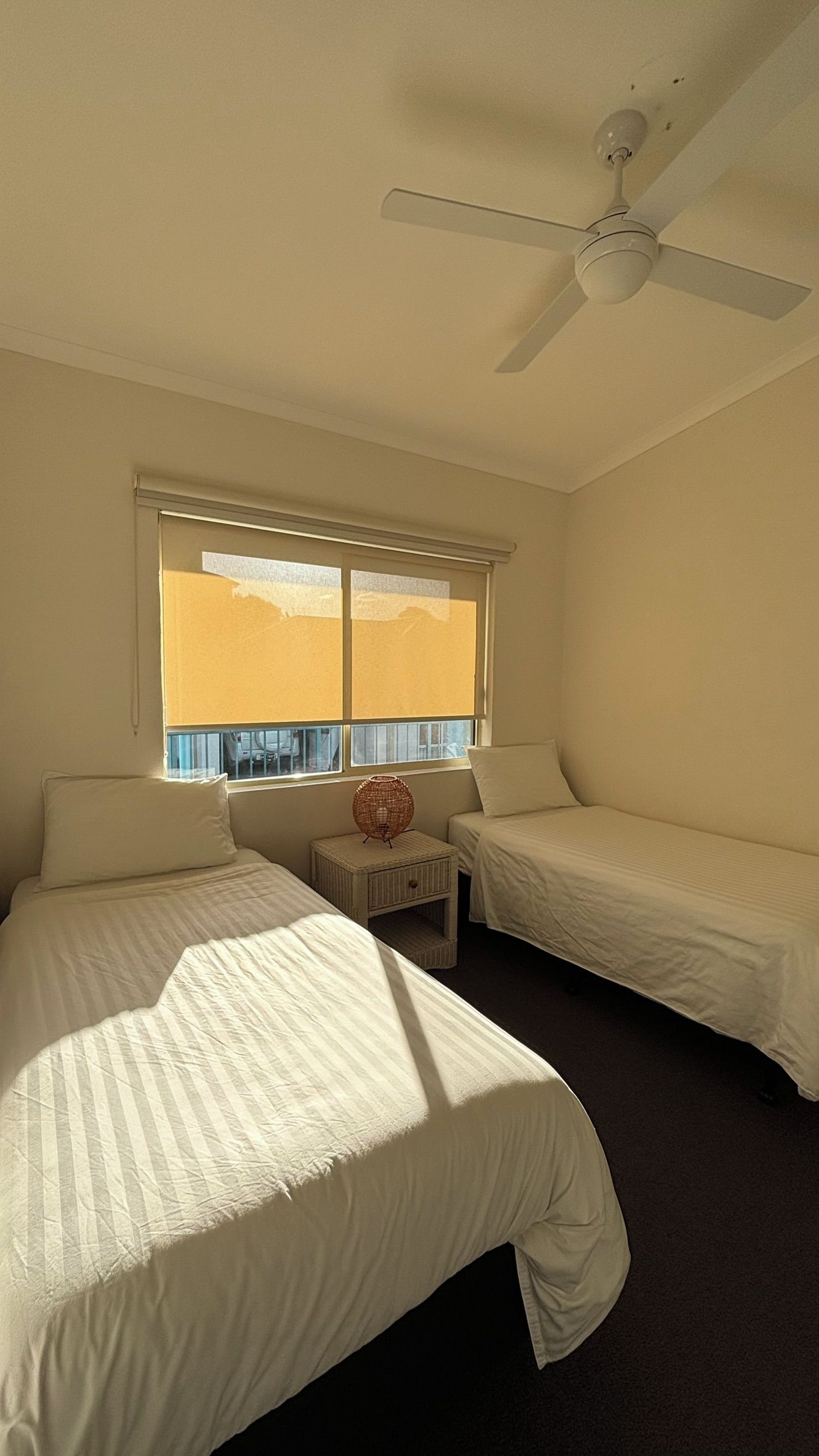 A room with two beds, a ceiling fan, a window with blinds, and a small table with a decorative item.