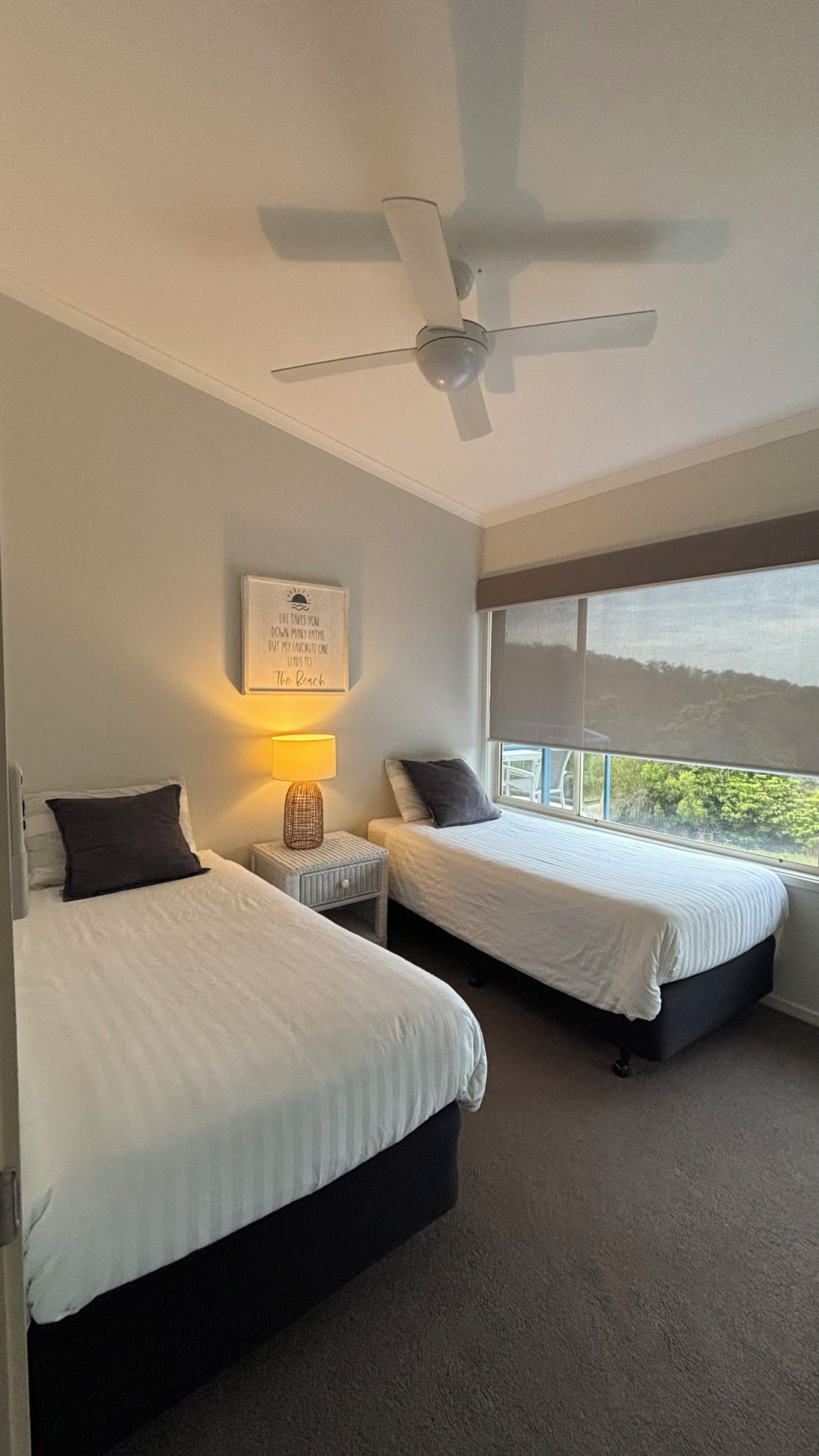 A hotel room with two beds, a ceiling fan, a bedside lamp, and a window with a view.