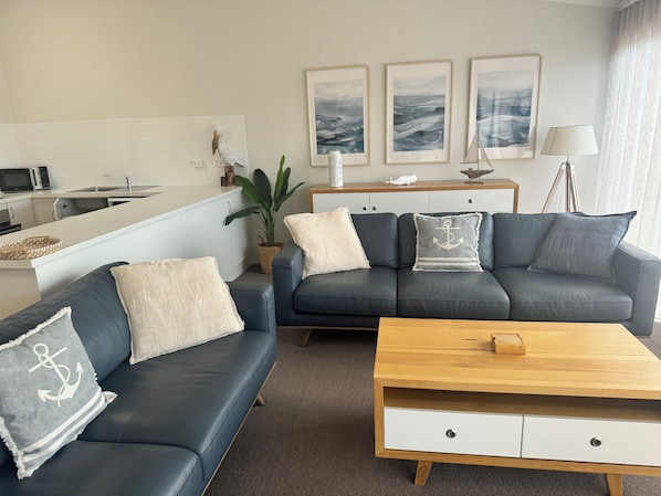 A modern living room with a dark grey sofa set, a wooden coffee table, and a white side table. There are three framed artworks on the wall, a potted plant, and a lamp on a side table. - Bluewater Apartments Merimbula (Merimbula)
