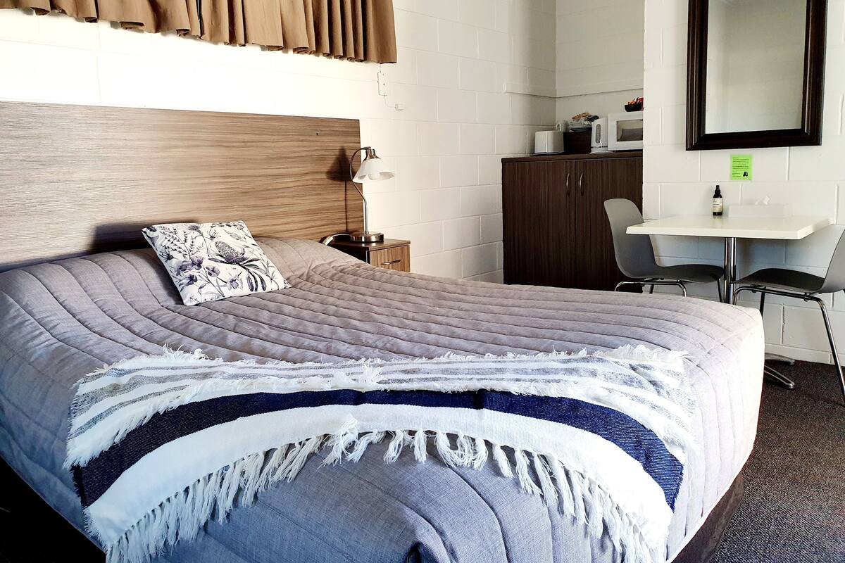 Una cama bien hecha con cabecera, una mesita de noche con lámpara, un escritorio con sillas y un espejo en la pared.