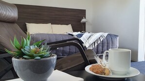 A neatly arranged hotel room with a bed, a bedside table with a cup and cookies, a potted plant, and a framed picture on the wall.