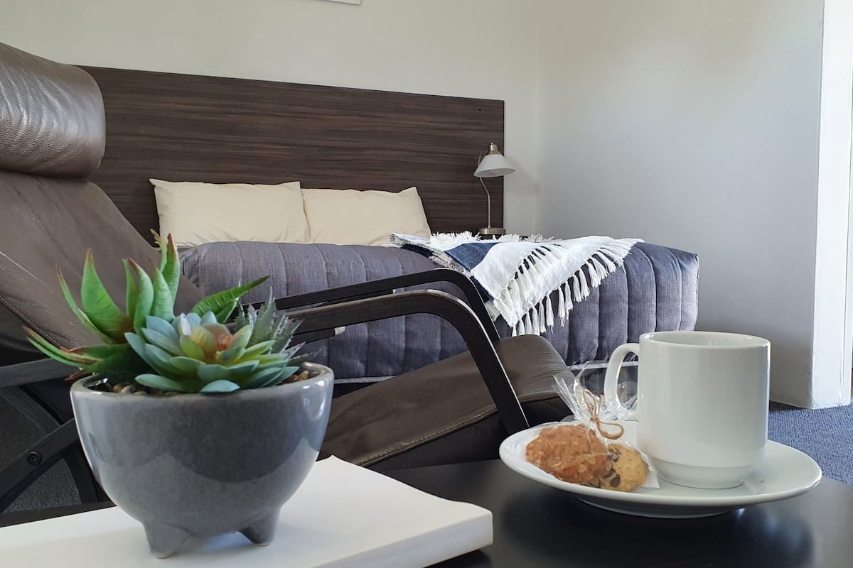 Habitación de hotel ordenada con una cama, una mesita de noche con una taza y galletas, una planta en maceta y un cuadro en la pared.