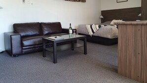 A modern, compact room with a brown leather sofa, a small table with a bottle and glasses, a bed with a grey blanket, and a framed picture on the wall.