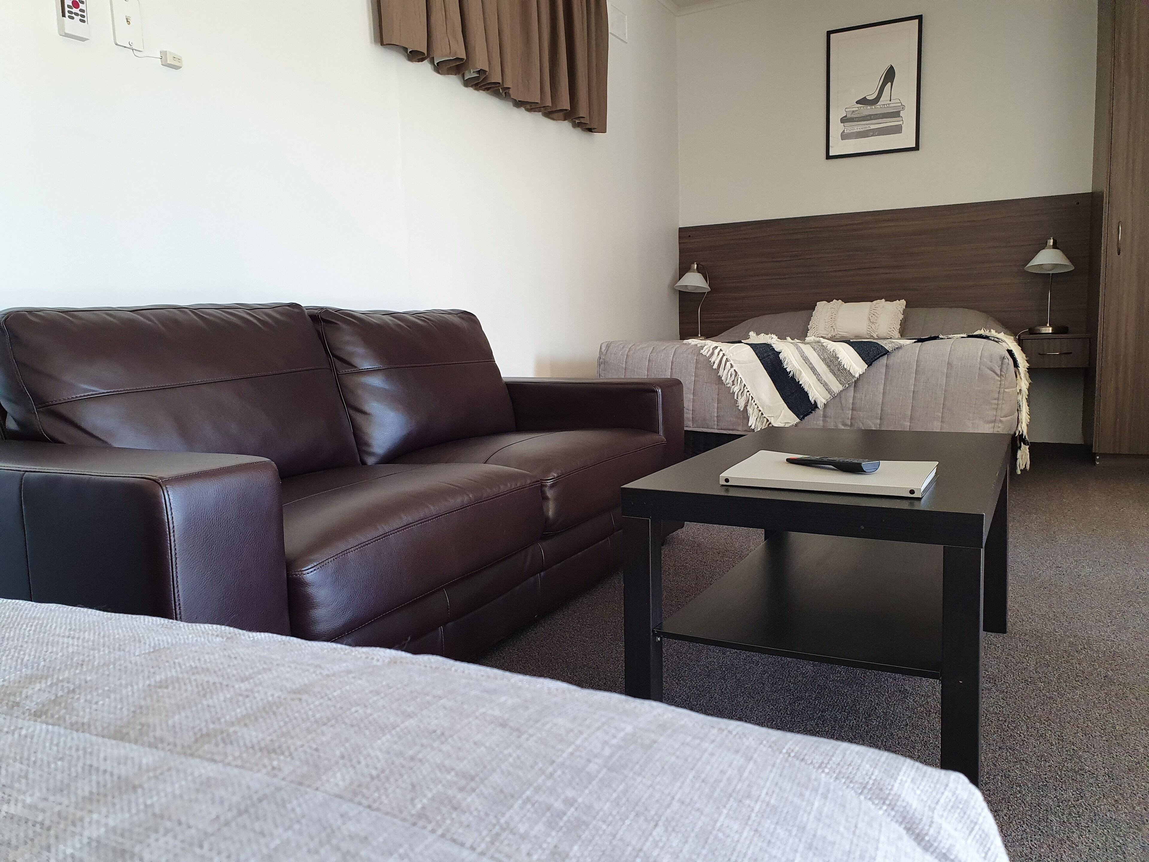 Habitación de hotel con un sofá de cuero marrón, una cama con una colcha a rayas grises y blancas, una mesita con un control remoto y un cuadro con tacones altos en la pared.