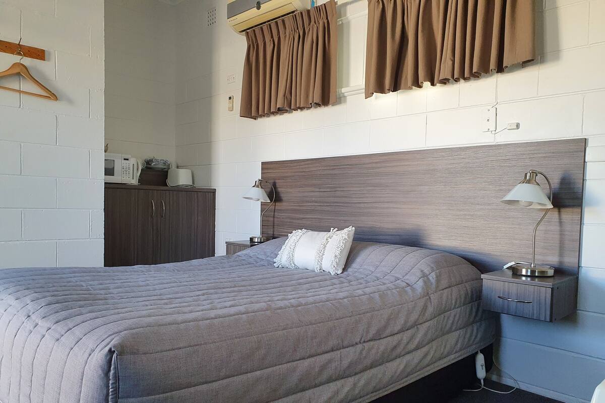 Una cama bien hecha con un edredón gris, un cabecero de madera, una lámpara de noche y una mesita de noche con una lámpara.