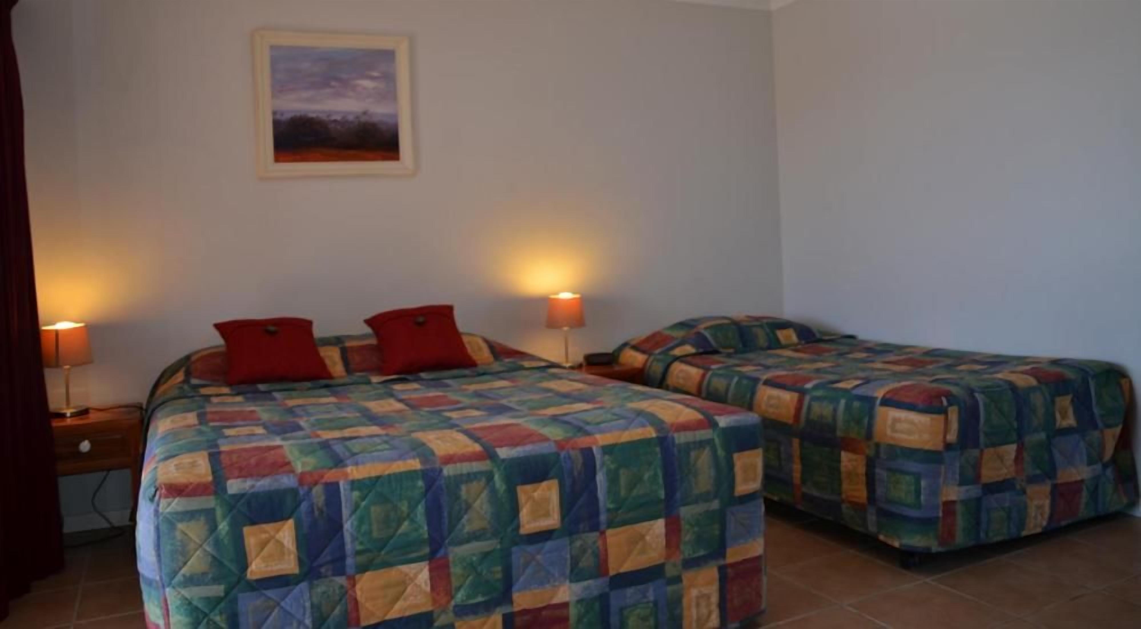 A room with two beds, each with a colorful quilt, a painting on the wall, and two bedside lamps.