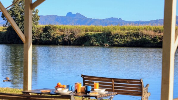 Una mesa con platos y tazas, un banco y una vista de un río con montañas al fondo.