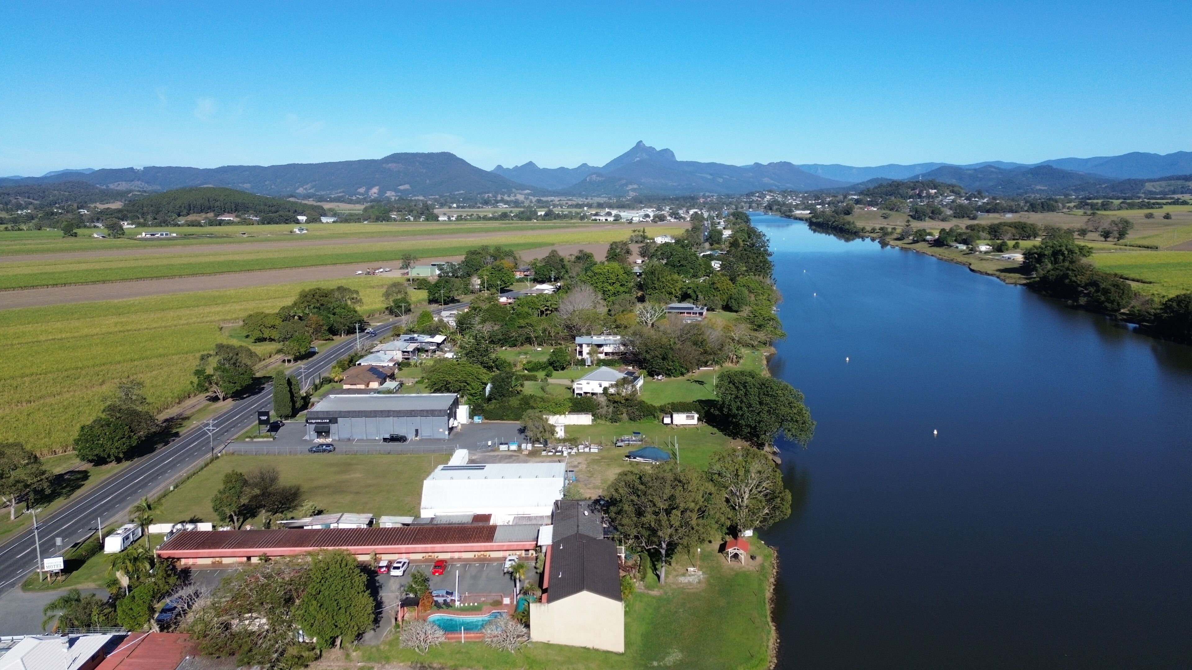 An aerial view of a riverside town with a mix of residential and commercial buildings, a road, and a swimming pool area.