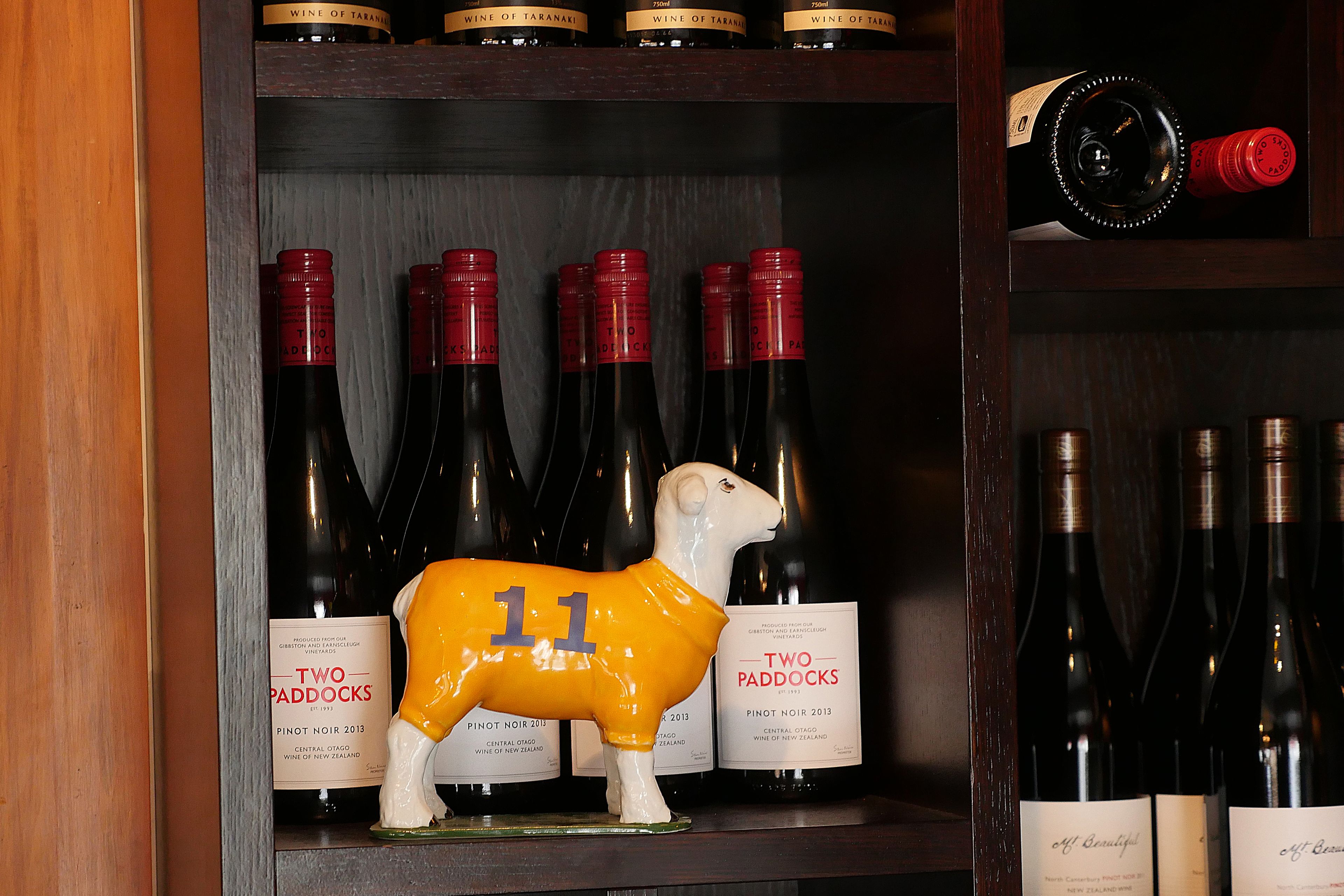 Una botellera con botellas de Two Paddocks Pinot Noir 2013, una figurita de oveja de cerámica blanca y una botella de vino tinto.