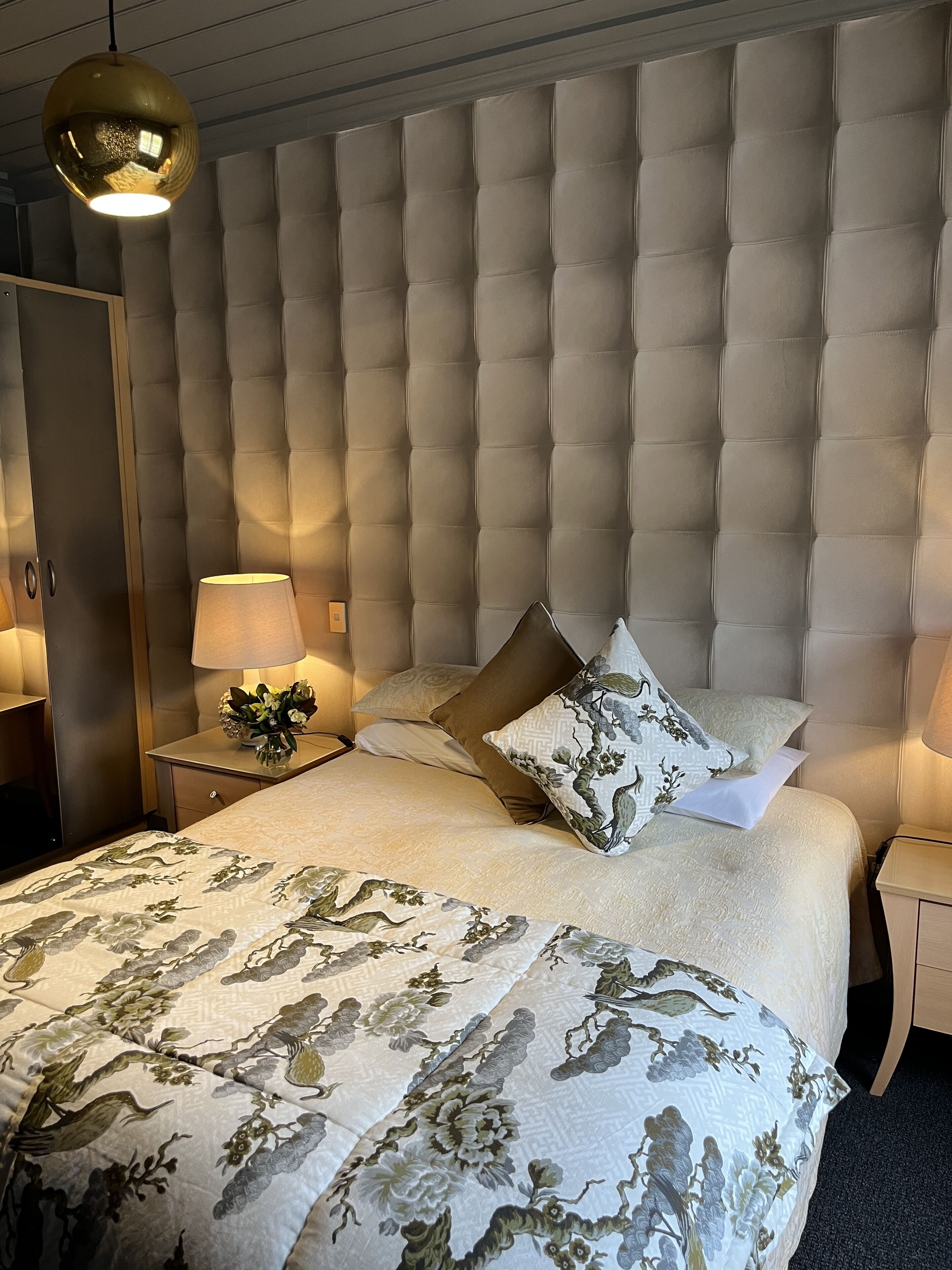A bedroom with a tufted headboard, a floral bedspread, two bedside lamps, and a nightstand with a vase of flowers.