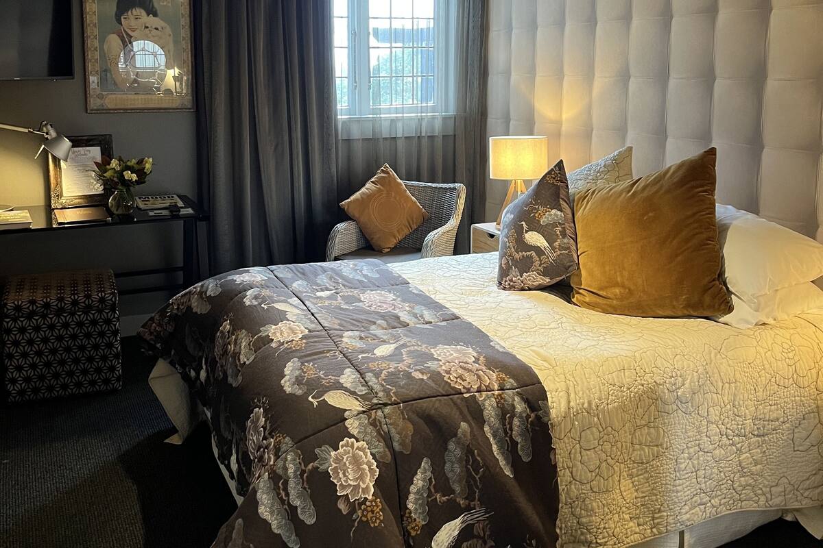 A hotel room with a large bed, a headboard, a desk with a lamp, a window with curtains, and a wall-mounted air conditioner.