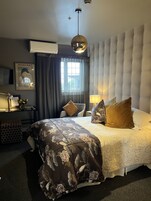 A hotel room with a large bed, a headboard, a desk with a lamp, a window with curtains, and a wall-mounted air conditioner.