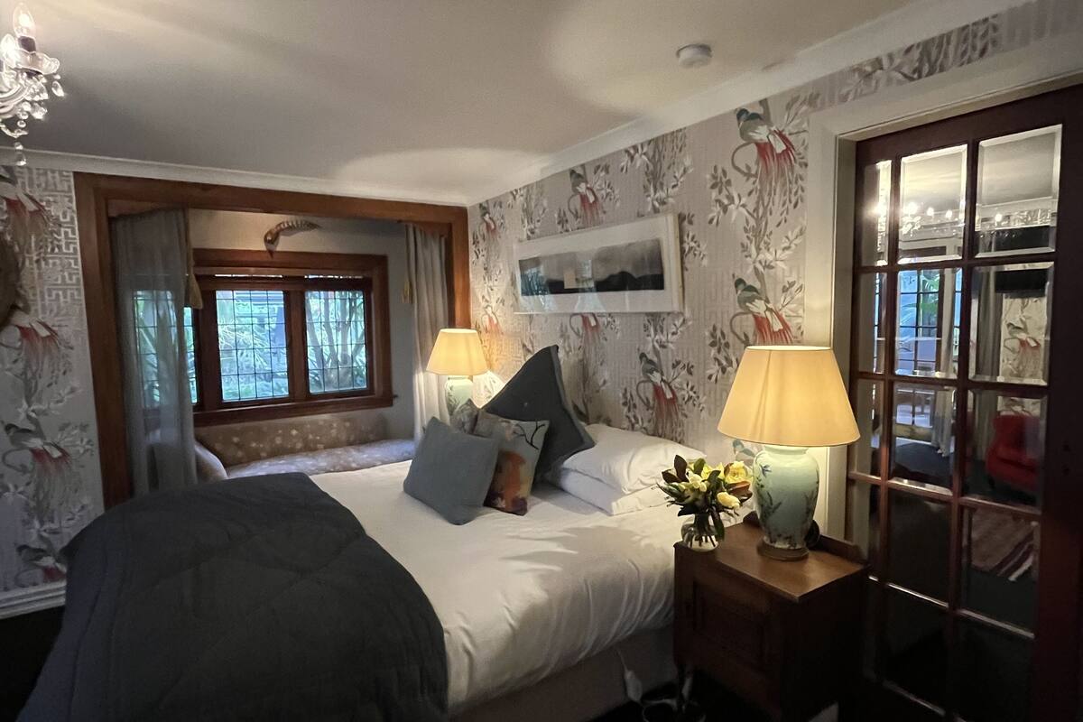 A bedroom with a bed, two bedside lamps, a window with curtains, and a wall with floral wallpaper.