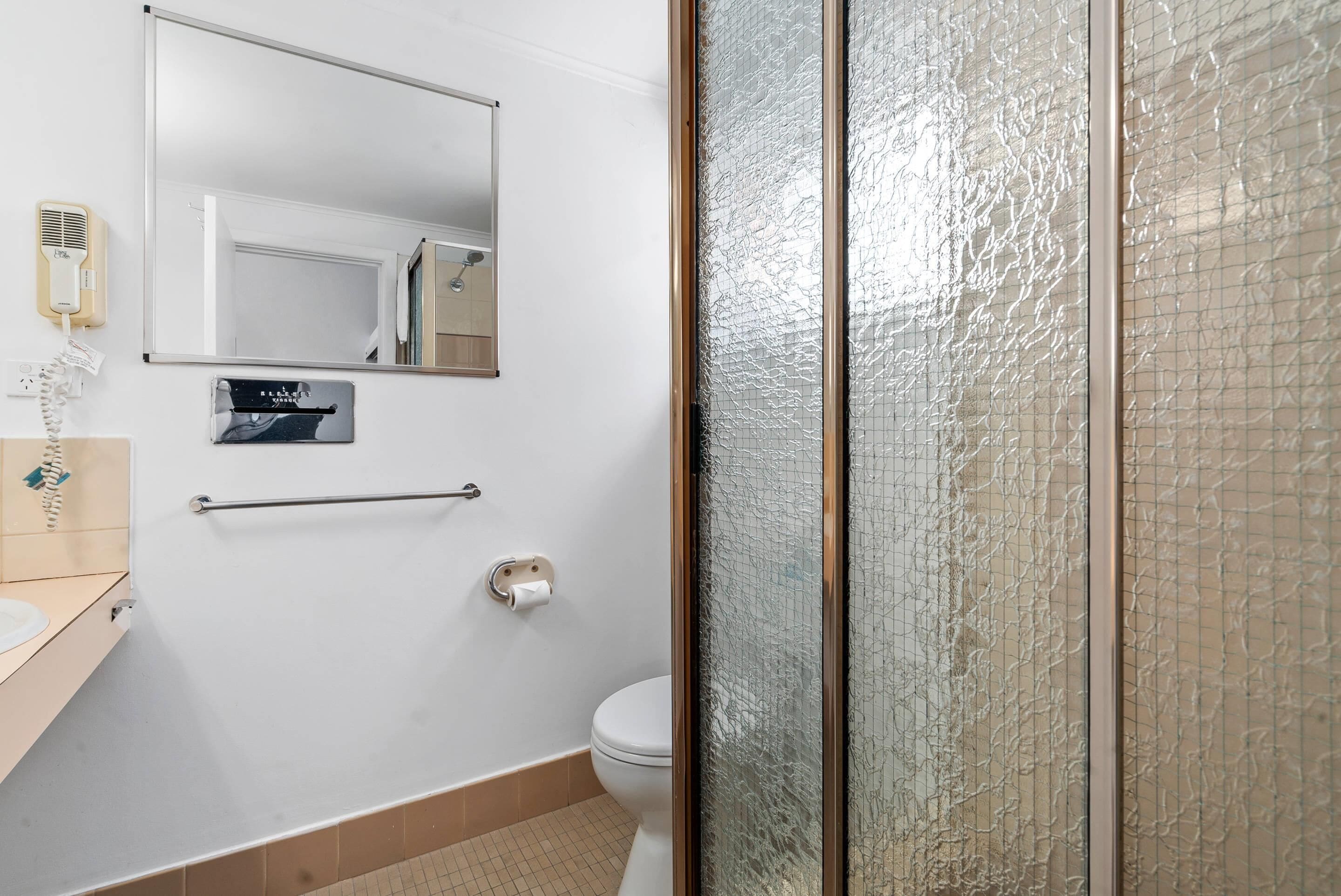Un baño con inodoro, secamanos, toallero, espejo y mampara de vidrio.