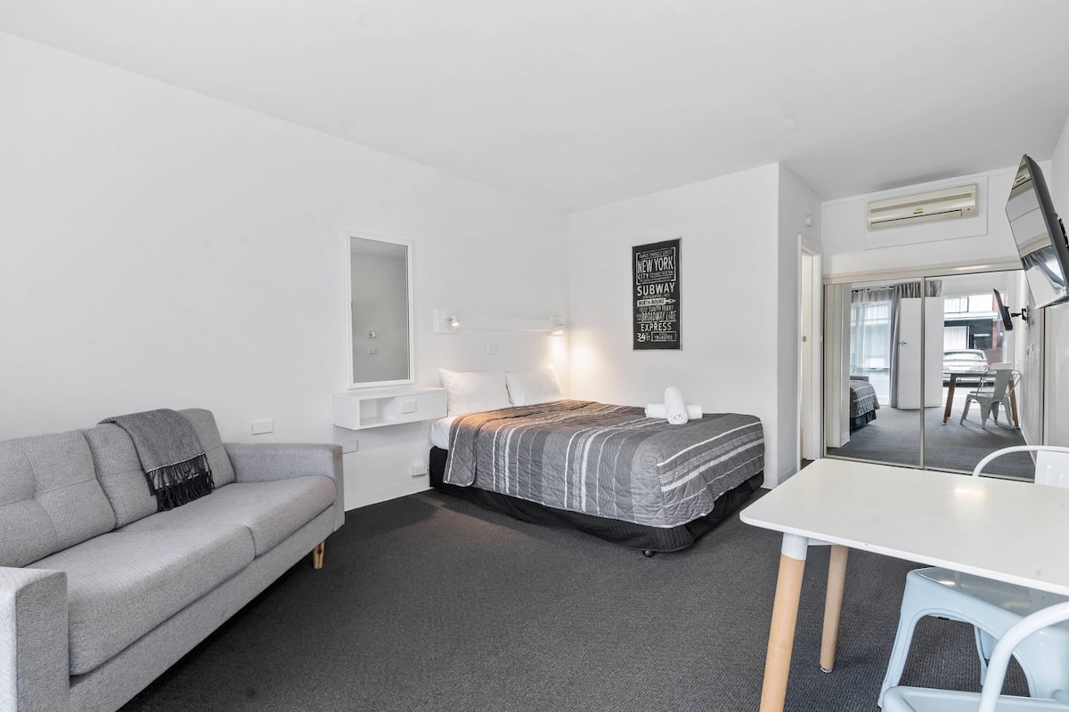 A modern hotel room with a gray sofa, a bed with a striped blanket, a white table, and a wall-mounted air conditioning unit.