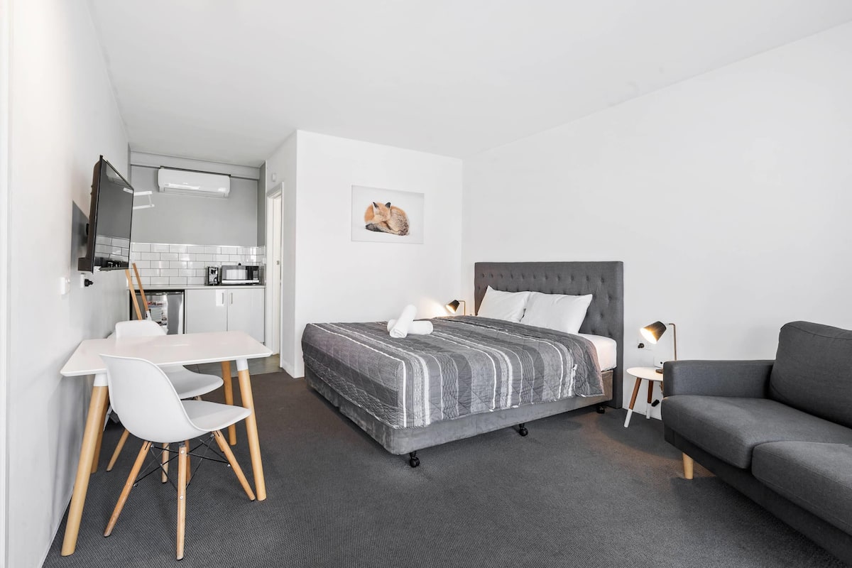 A modern hotel room with a bed, a sofa, a dining table with chairs, a kitchen area, and a flat-screen TV.