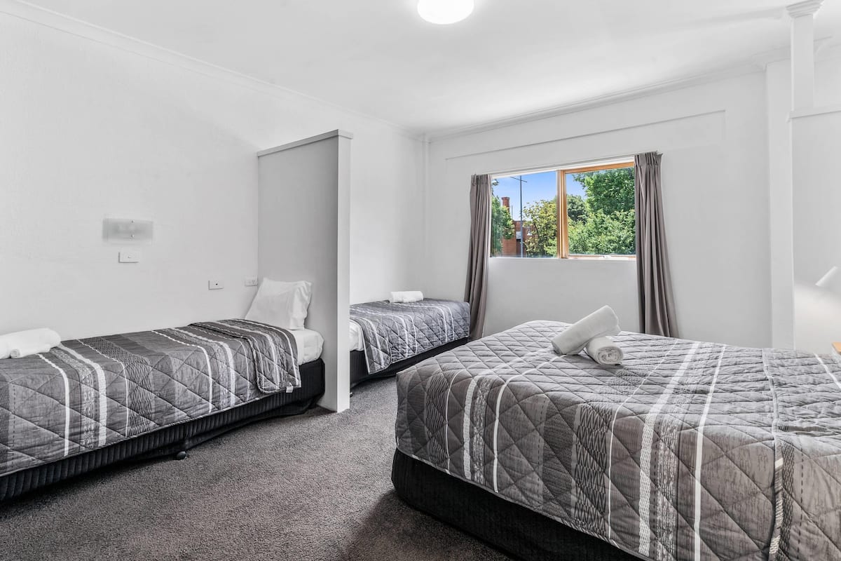 A hotel room with two single beds, a window with curtains, and a view of greenery outside.