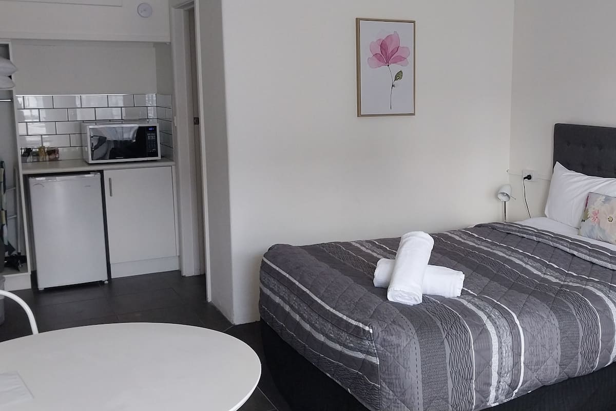 A hotel room with a bed, a small table, a chair, a microwave, and a refrigerator.