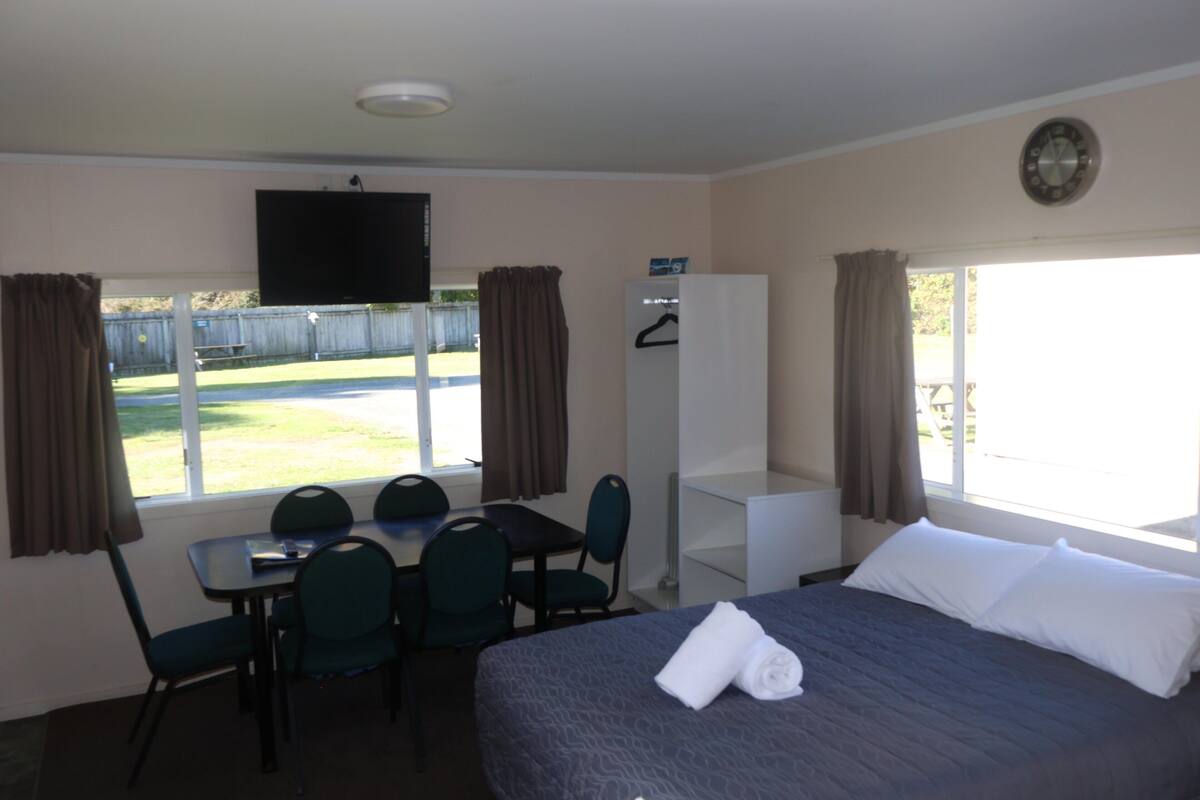 A hotel room with a bed, a desk with chairs, a television, and a view of a fenced yard.