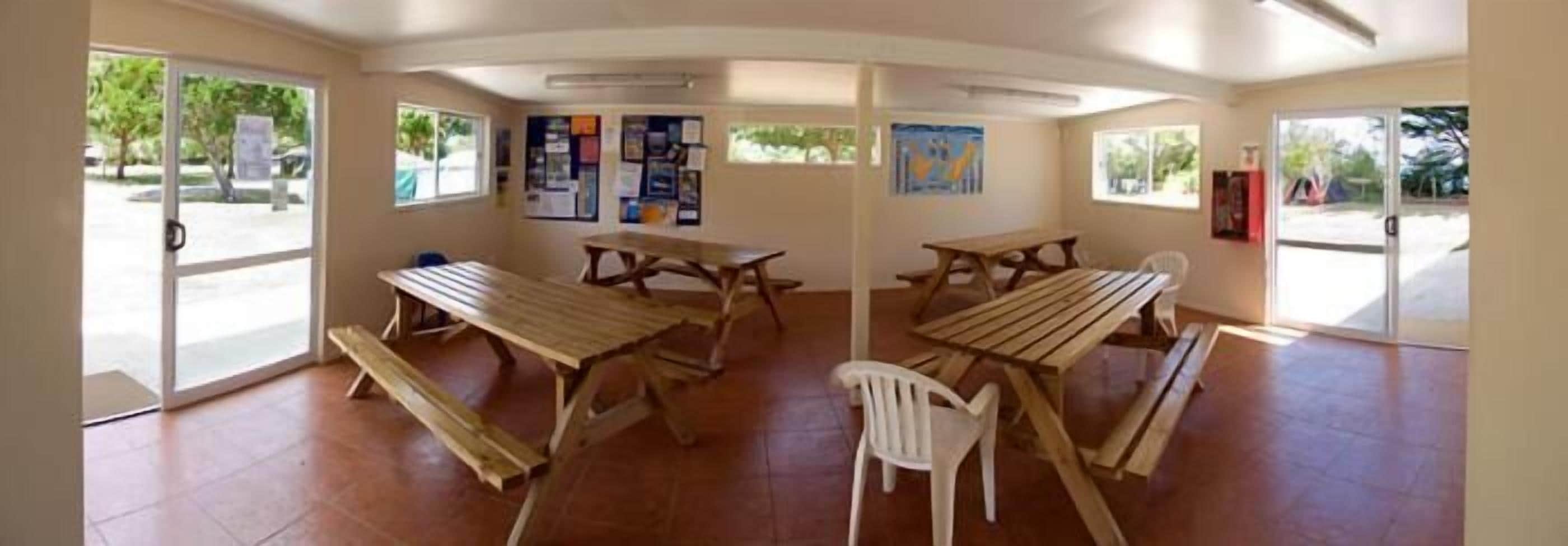 A room with a large table and benches, a white plastic chair, and a bulletin board on the wall.