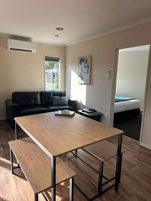 A modern, compact living space with a wooden dining table, a black leather sofa, and a bed in the adjacent room. - Pohara Beach Top 10 Holiday (Pohara)