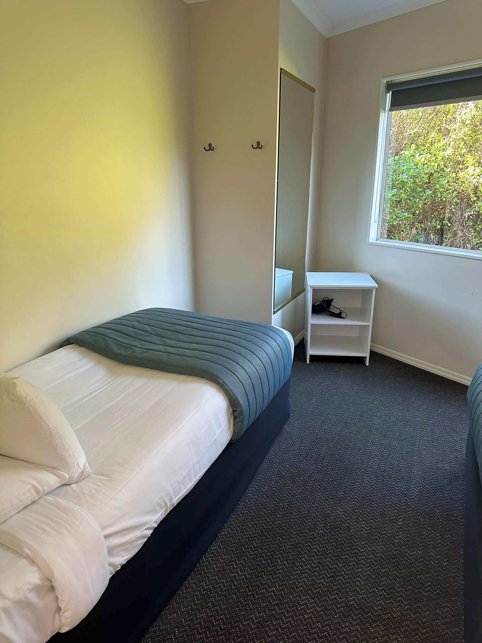A bedroom with a bed, a small white nightstand, a mirror, and a window with a view of trees.