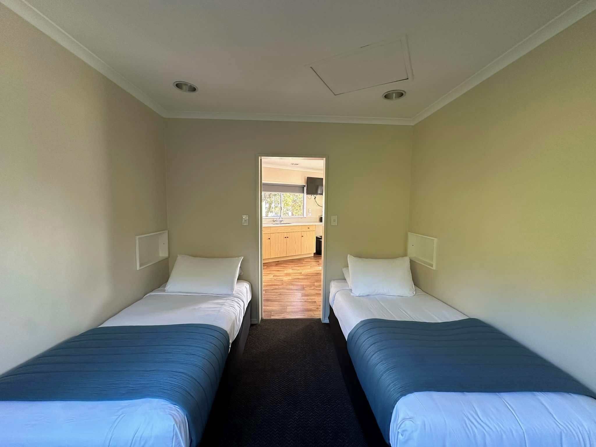 Een kamer met twee eenpersoonsbedden, een klein raam en een deur naar een keuken.