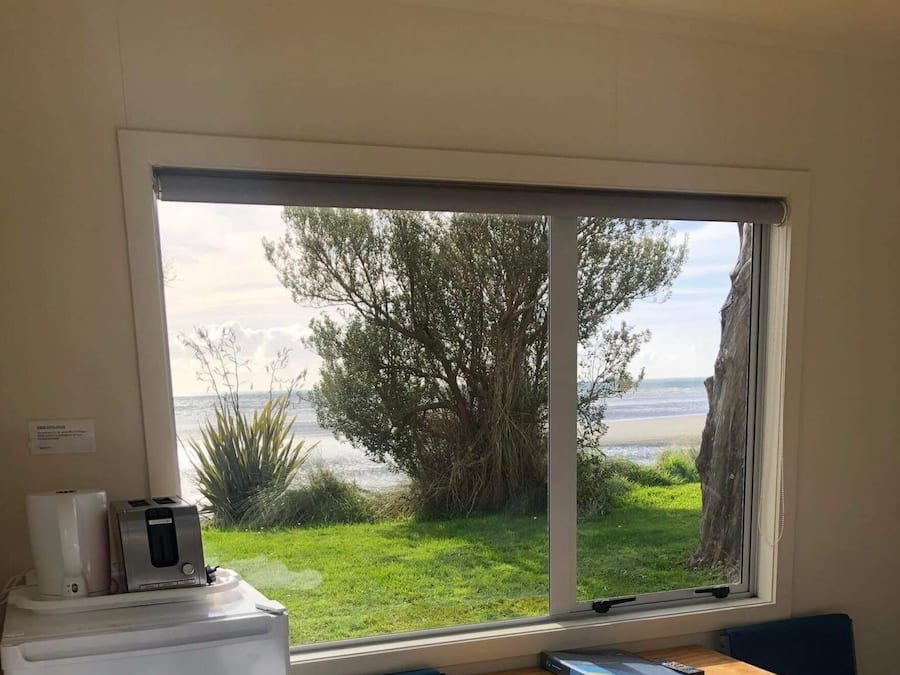 Una habitación con una cama, un escritorio con dos sillas, una kitchenette pequeña y una ventana con vista a la playa y árboles.
