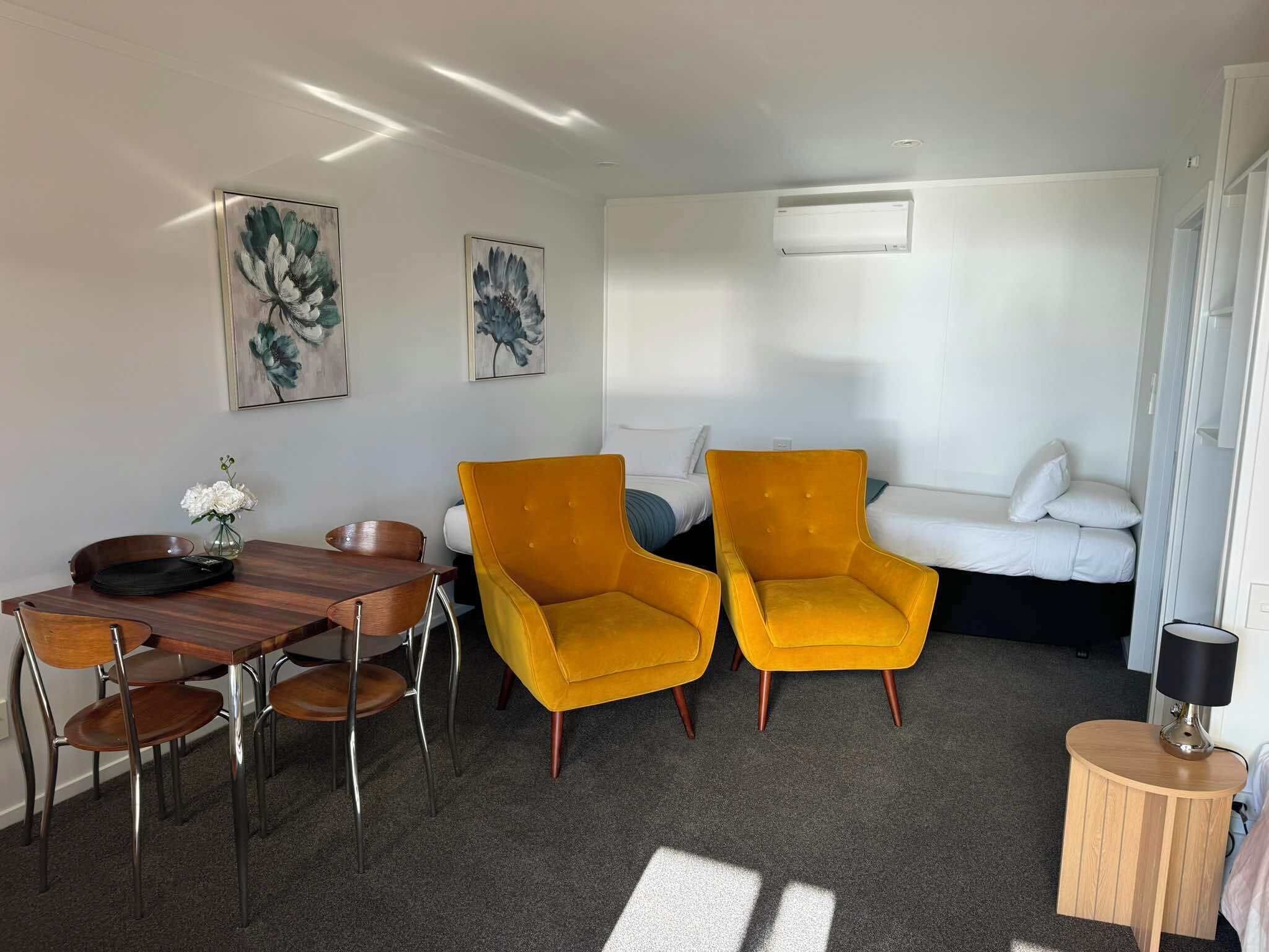Moderni hotellihuone, jossa on puinen ruokapöytä, kaksi keltaista nojatuolia, valkoinen sohva ja pieni pyöreä puinen sivupöytä.