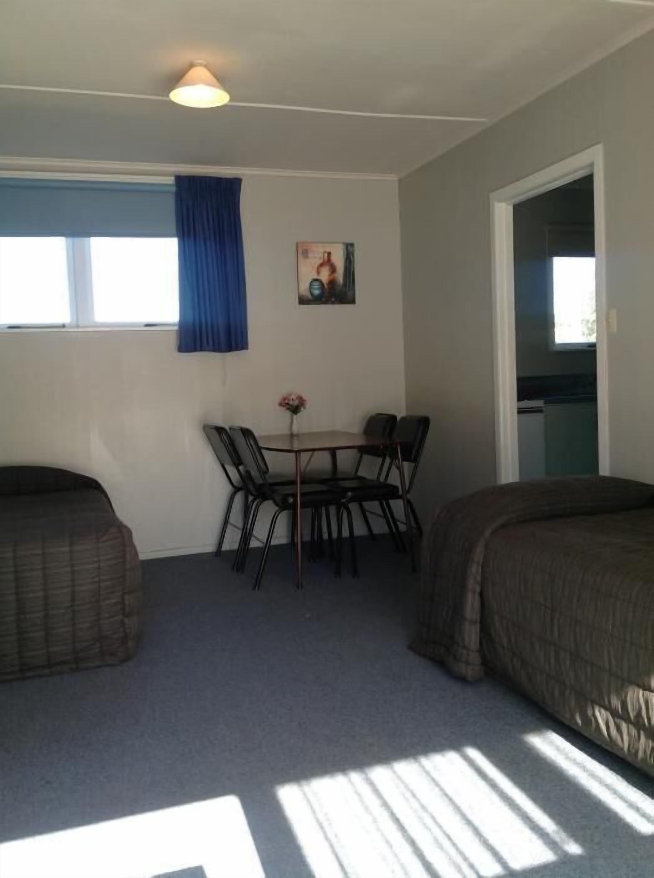 A room with two beds, a dining table with chairs, a painting on the wall, and a window with blue curtains.