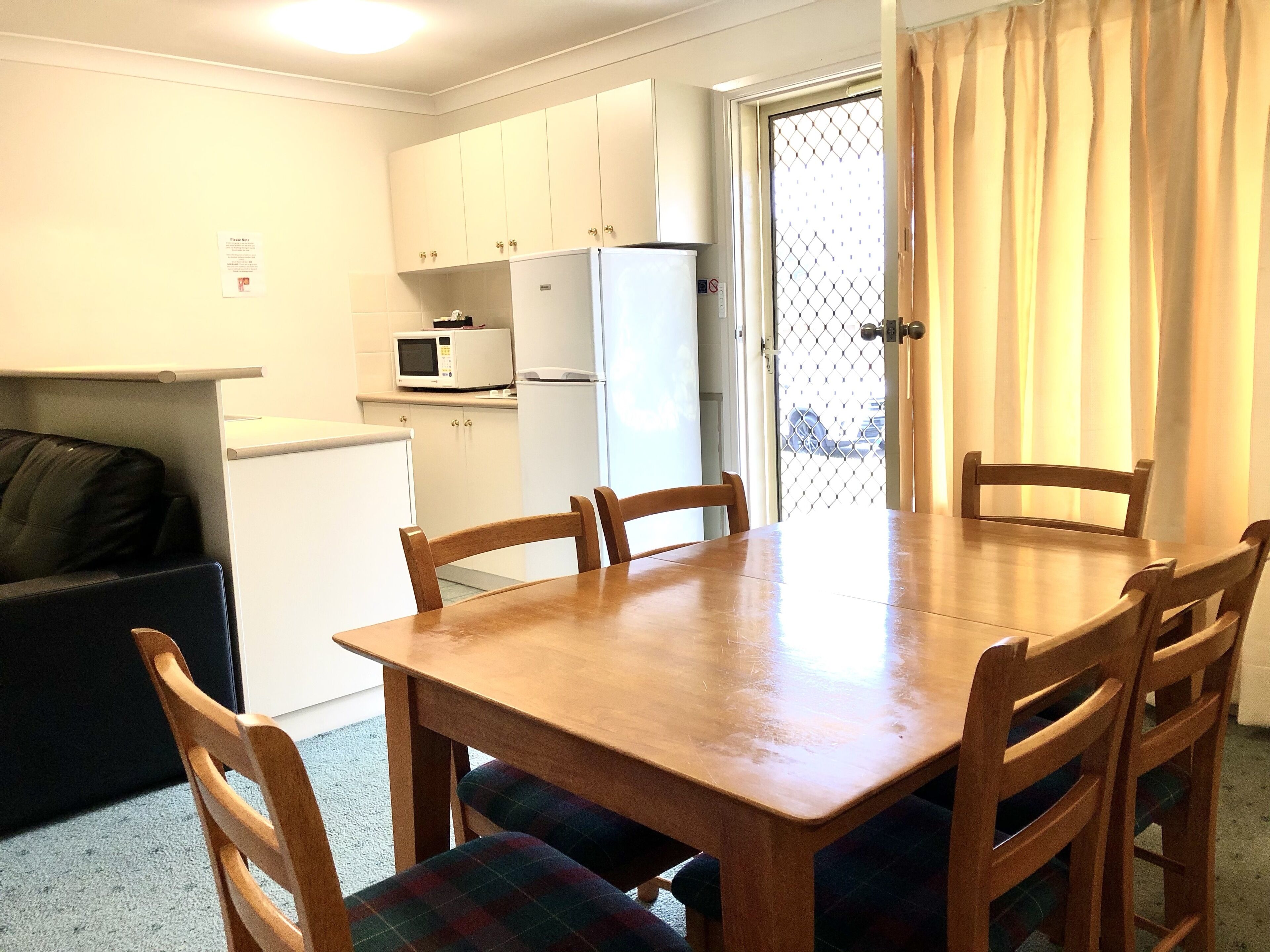 A compact kitchen with white cabinets, a microwave, and a refrigerator. There is a dining table with four chairs and a sofa in the corner.