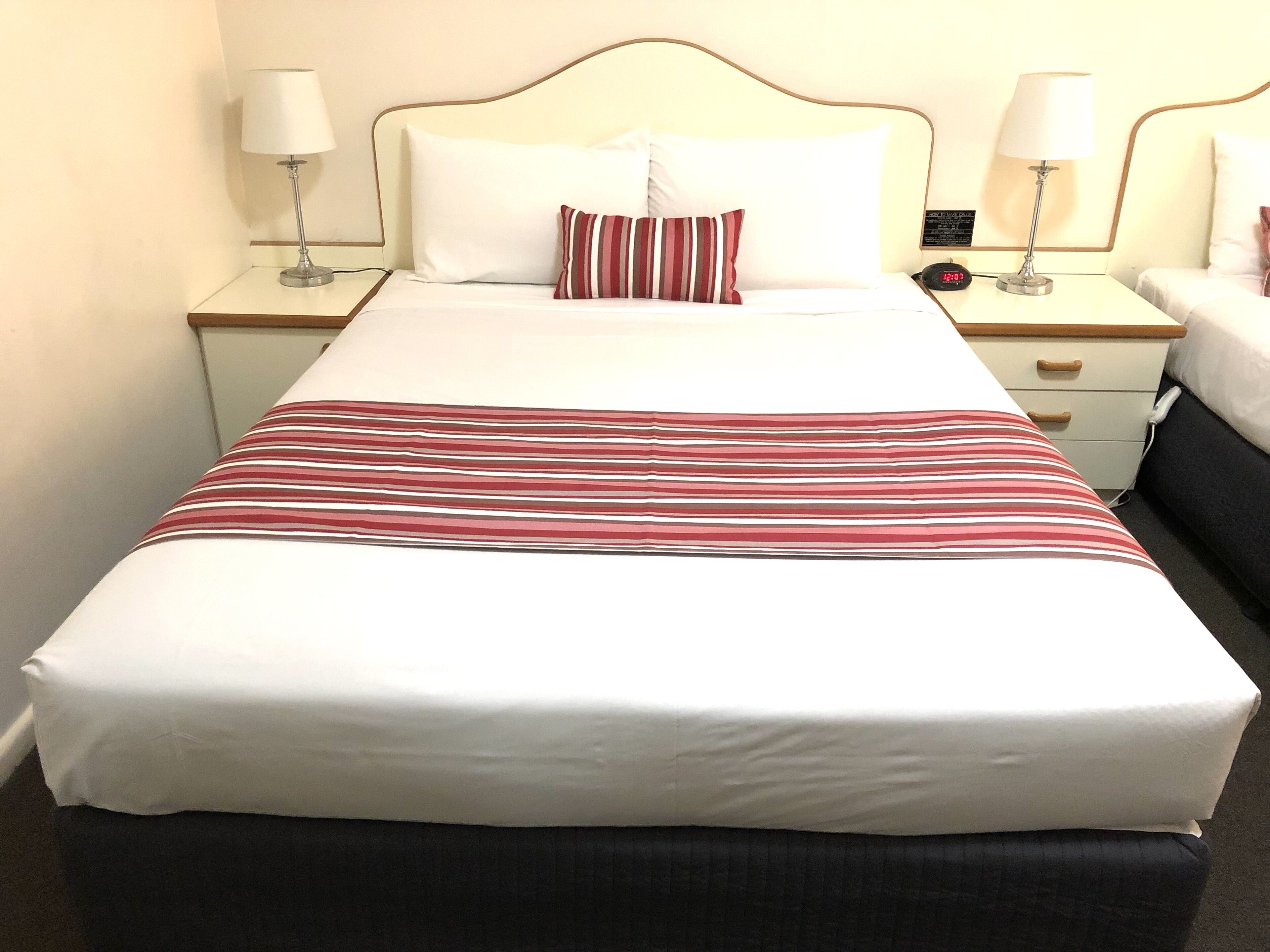 A hotel room with a large bed, two bedside tables with lamps, a nightstand with a clock, and a dark-colored bench at the foot of the bed.