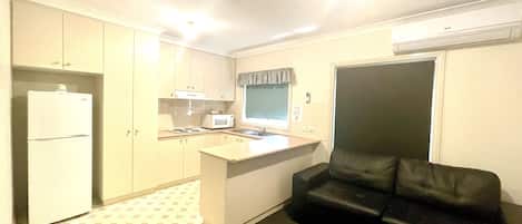 A compact kitchen with a refrigerator, microwave, and stove. There is a black leather couch and a window with a curtain.