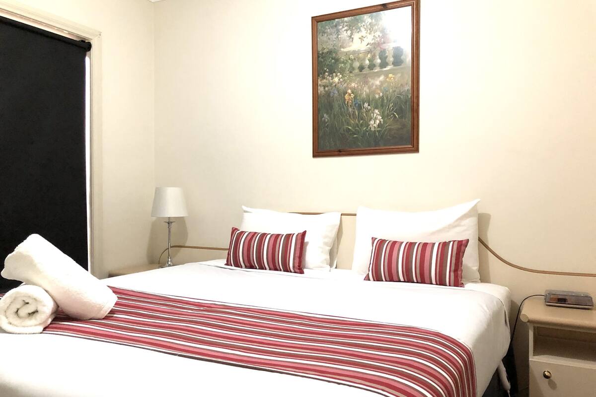 A hotel room with a bed, a nightstand, a lamp, a painting, and a window with a black blind.