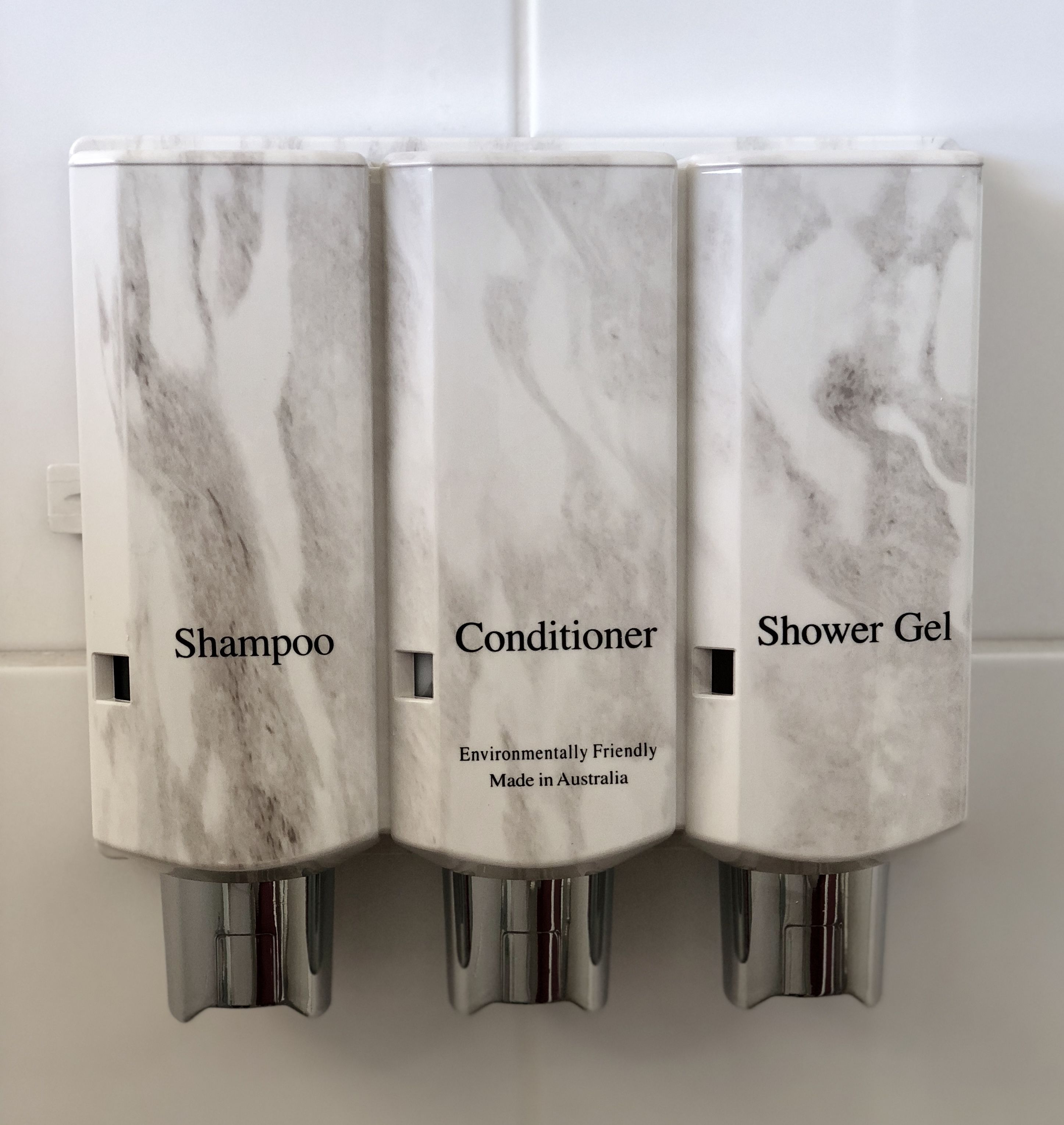 Dispensador montado en la pared con tres compartimentos etiquetados 'Champú', 'Acondicionador' y 'Gel de ducha'.
