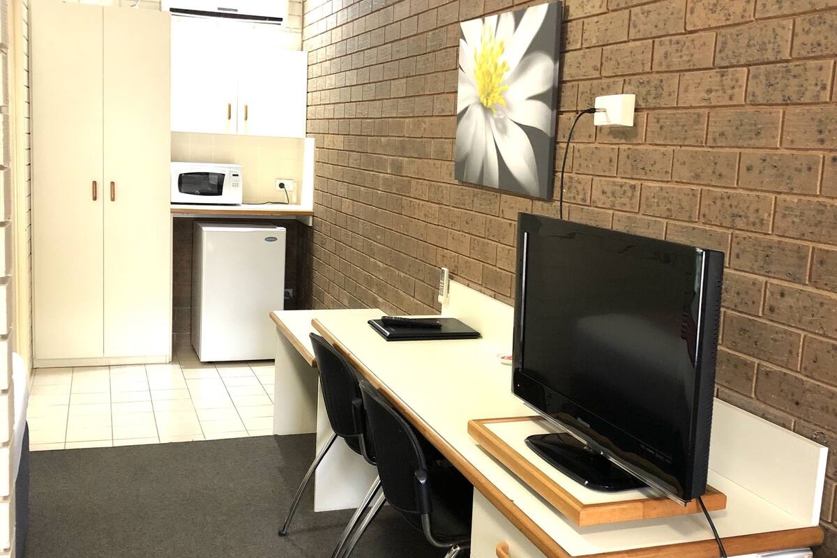 A compact kitchen with white cabinets, a microwave, and a refrigerator. A TV is mounted on a wooden desk with a black chair. A brick wall with a flower painting is visible.