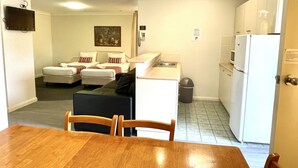 A compact hotel room with a kitchenette, a dining area with a wooden table and chairs, a small sofa set, and a trash bin.