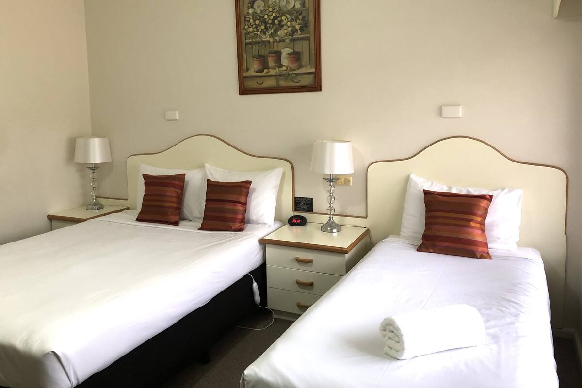 A hotel room with two beds, each with white linens and striped pillows. There are bedside tables with lamps and a digital clock. A framed artwork hangs on the wall above the beds.