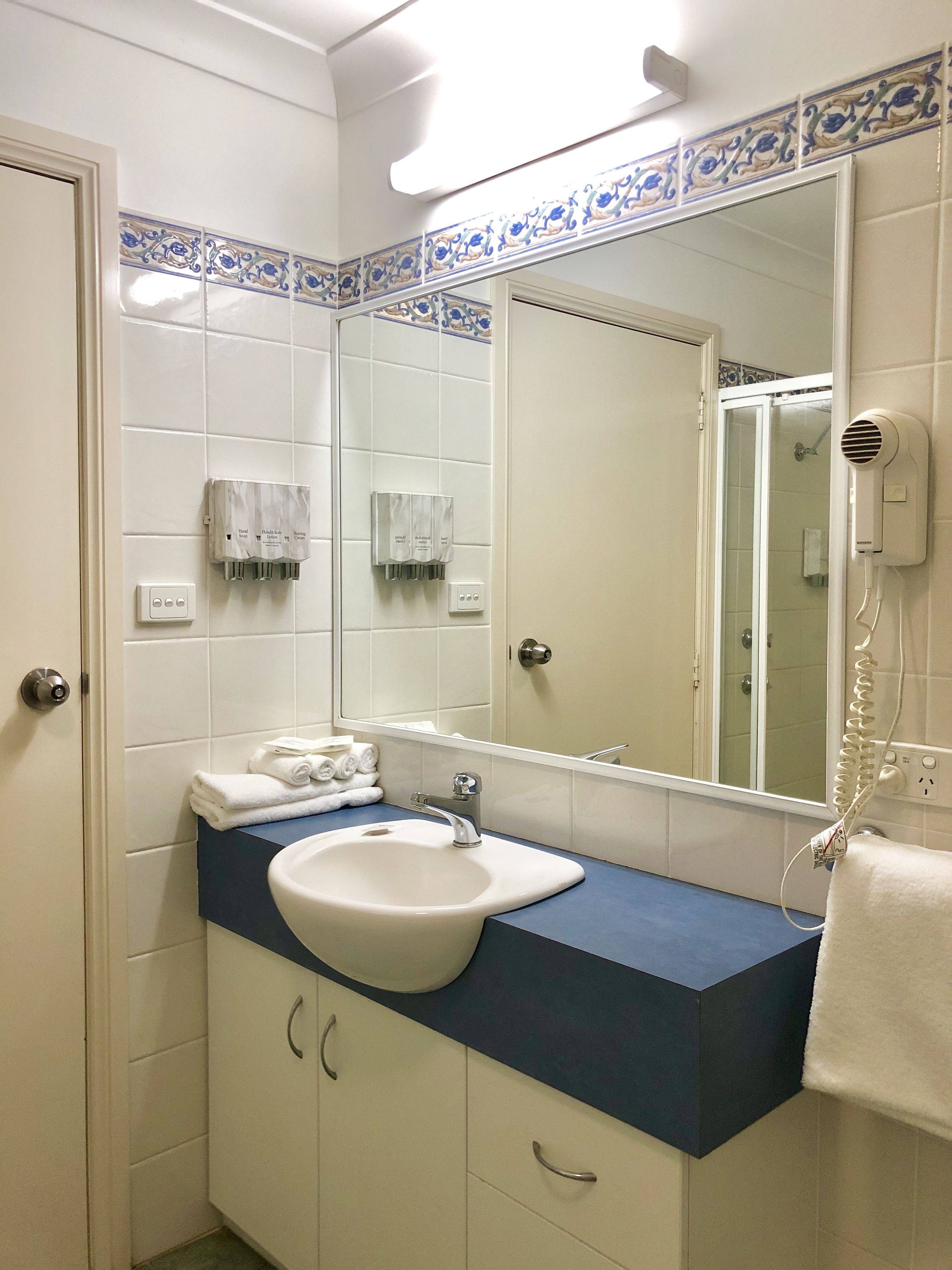 Une salle de bain avec un lavabo blanc, un plan de toilette bleu et un grand miroir. On y trouve des distributeurs de savon liquide et un sèche-cheveux fixés au mur.