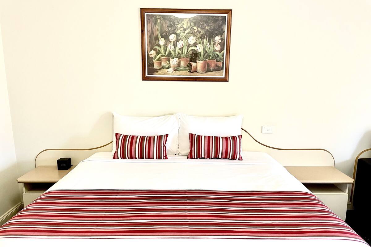 A hotel room with a bed, two pillows, a nightstand, a bedside table, and a framed picture on the wall.