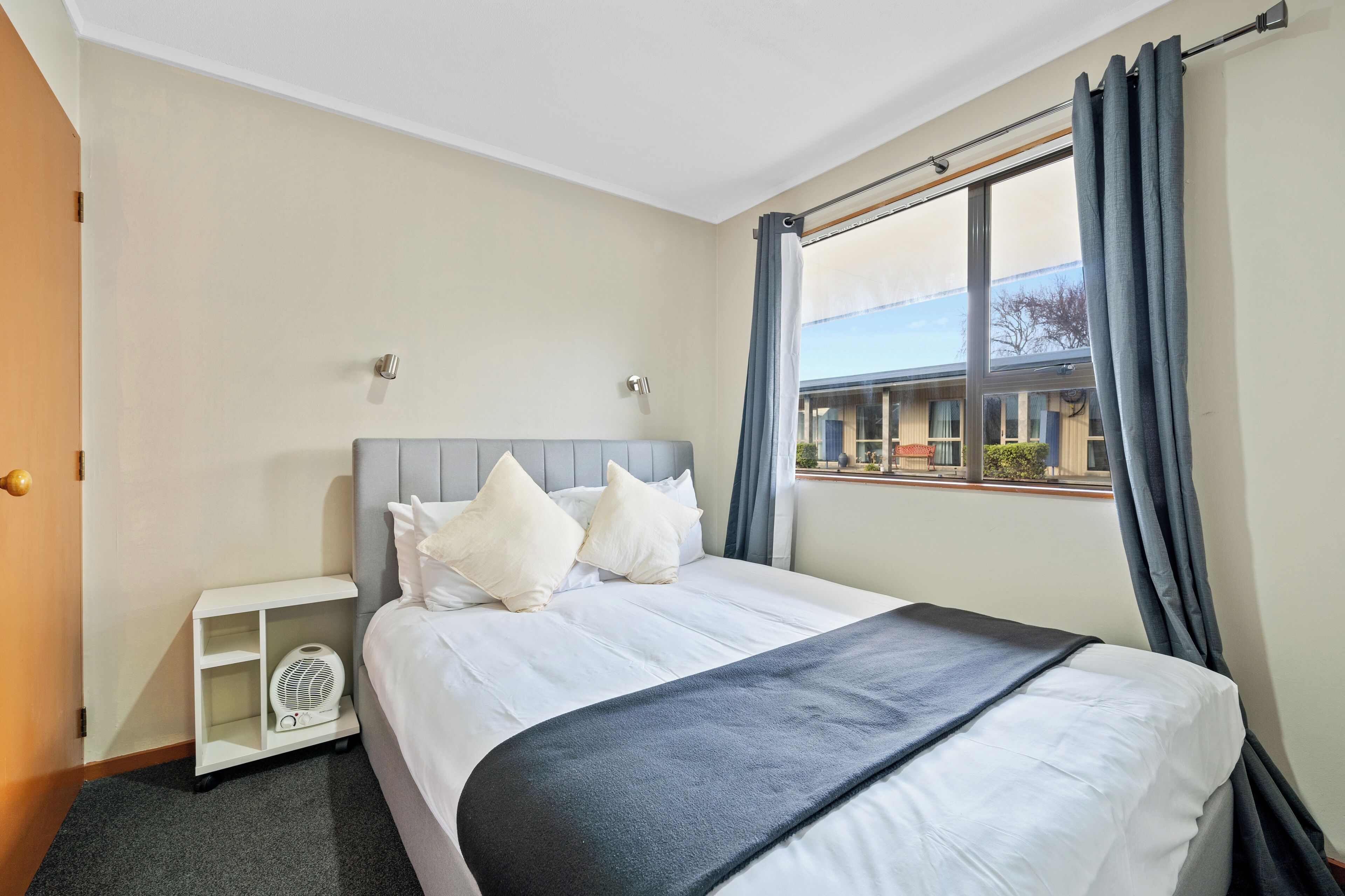 Une chambre d’hôtel avec un lit, un petit ventilateur blanc et une fenêtre avec des rideaux.