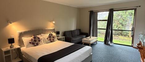 A hotel room with a large bed, a sofa, a small table, and a view of the outdoors through a large window.
