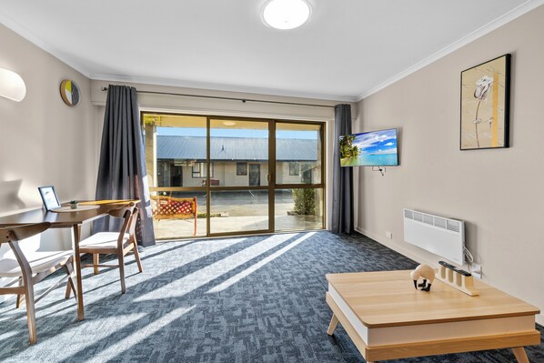 Habitación de hotel con un amplio ventanal corredero al exterior, un escritorio de madera con una silla, un televisor montado en la pared y un radiador.